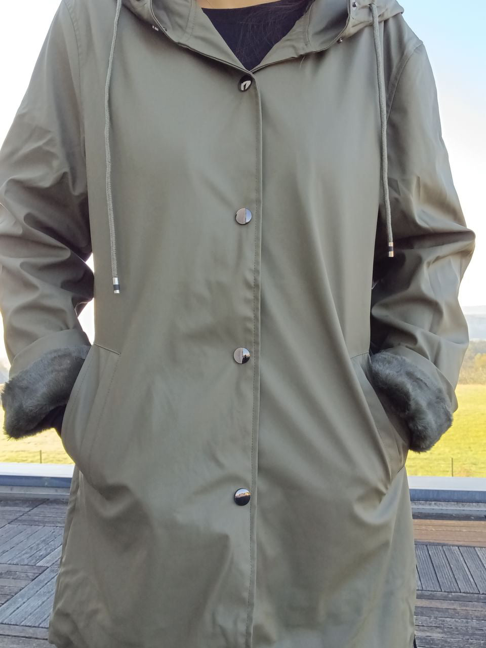Olive green Laura Jo raincoat with hood and faux fur cuffs, outdoors by Le Petit Placard