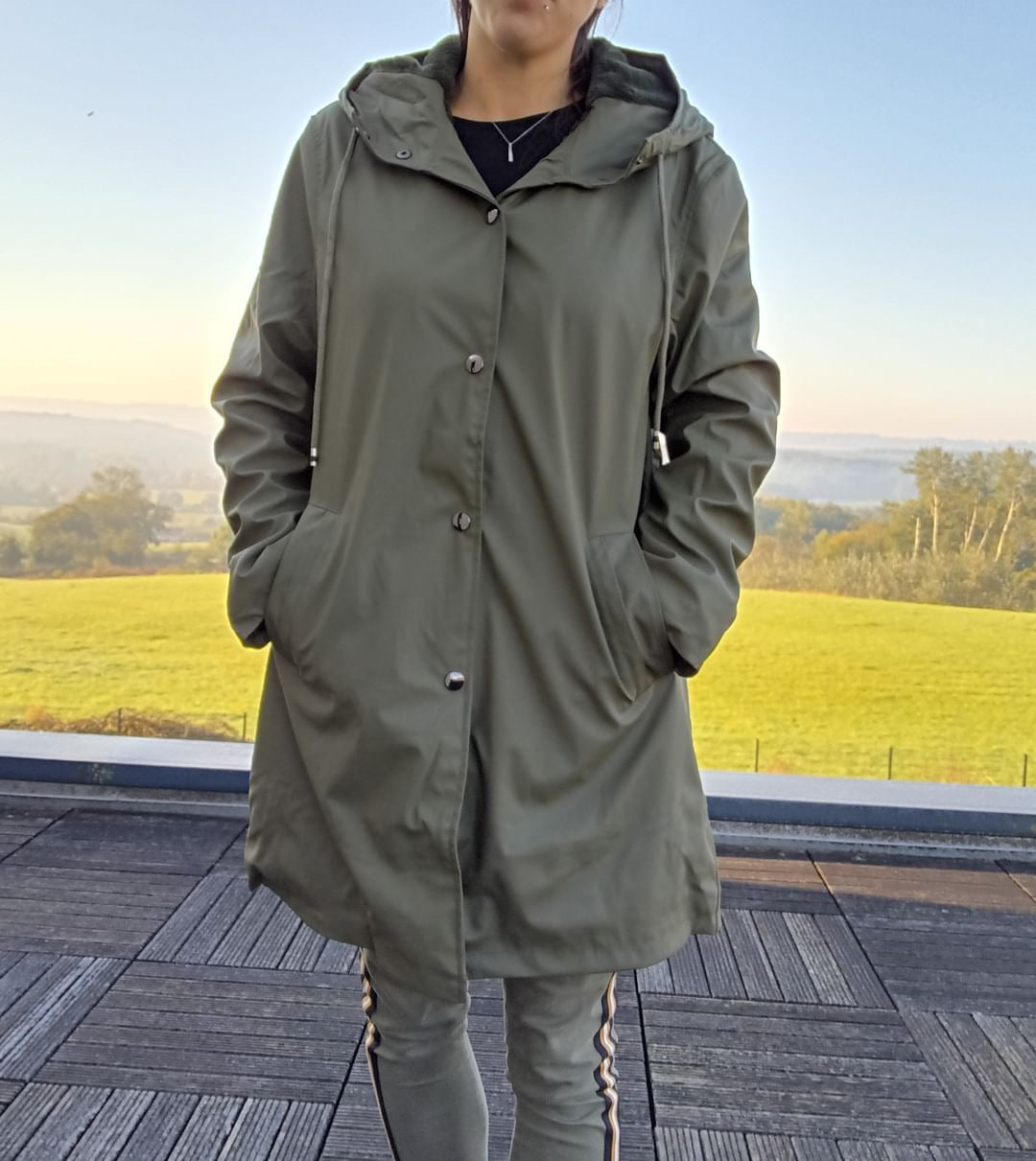 Woman wearing an olive Laura Jo raincoat on a terrace with green fields in the background