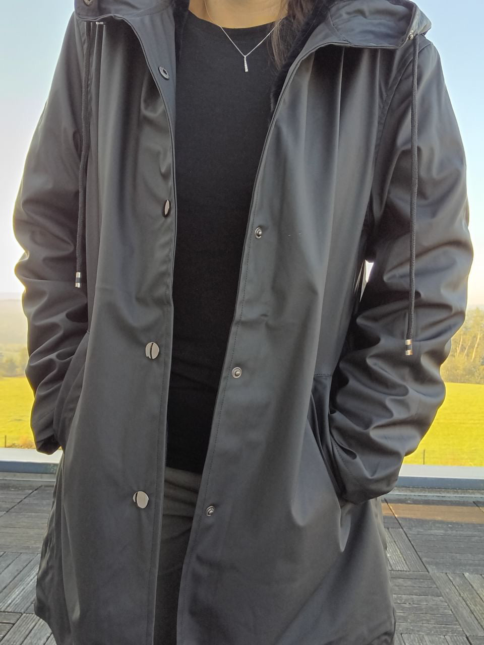 Black long waterproof raincoat with hood from Le Petit Placard, worn outdoors.
