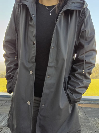 Black long waterproof raincoat with hood from Le Petit Placard, worn outdoors.