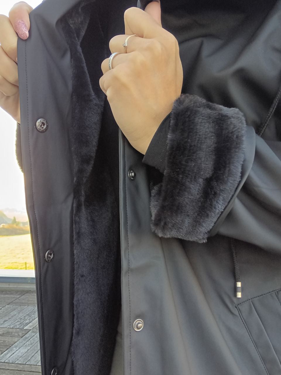 Black long waterproof coat with faux fur lining, shown open by a hand, Le Petit Placard