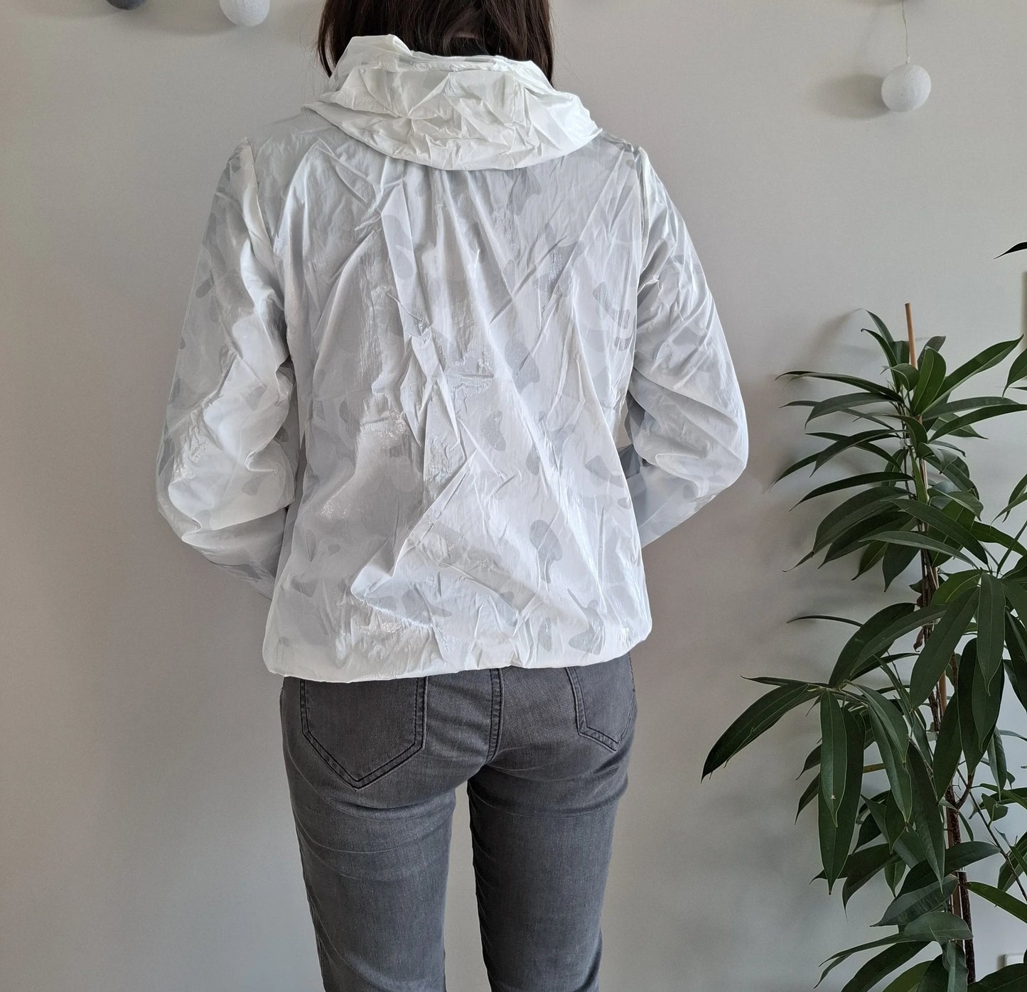 Back view of woman in white Laura Jo raincoat, gray jeans, near plant, Le Petit Placard