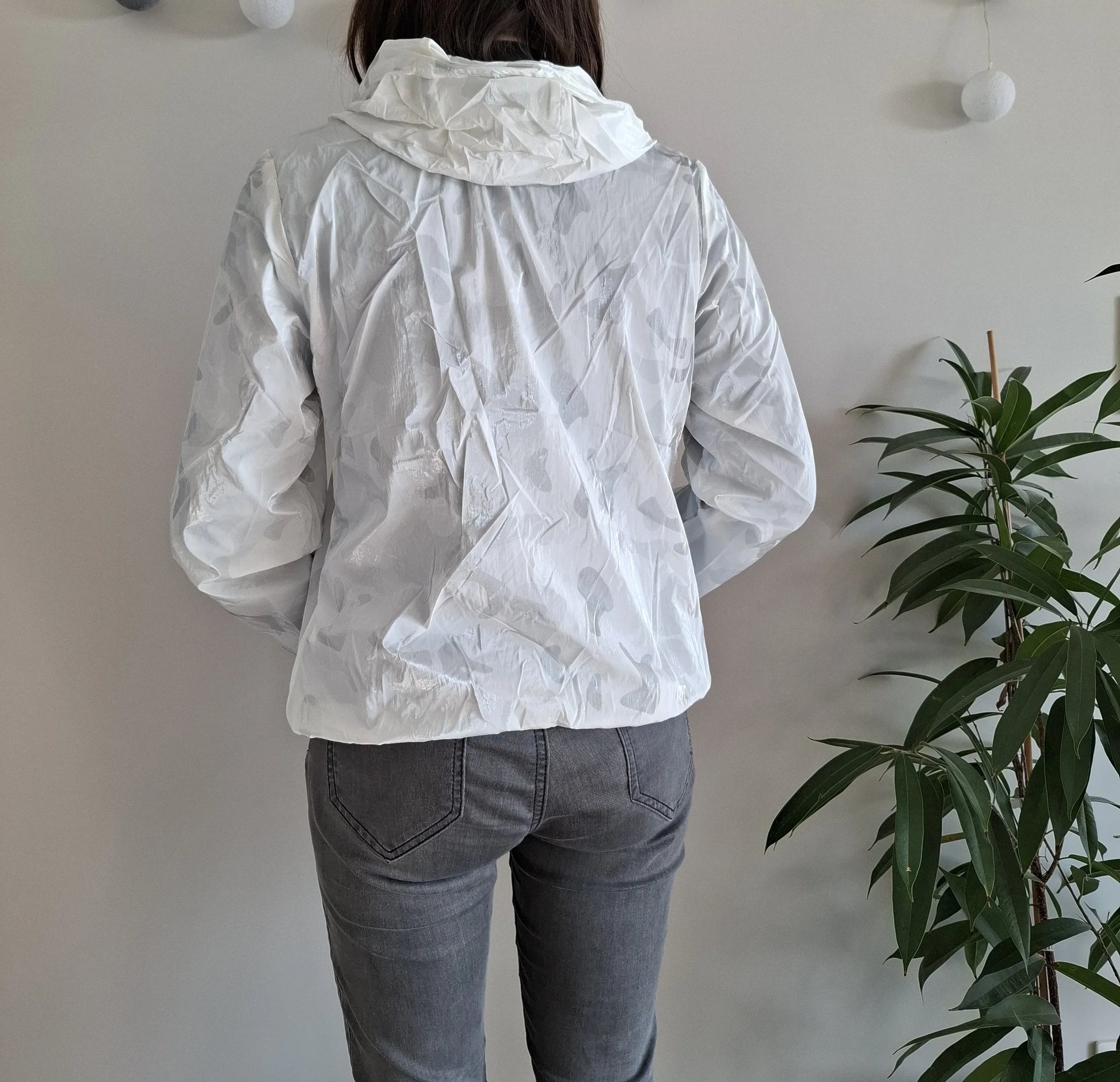 Back view of woman in white Laura Jo raincoat, gray jeans, near plant, Le Petit Placard