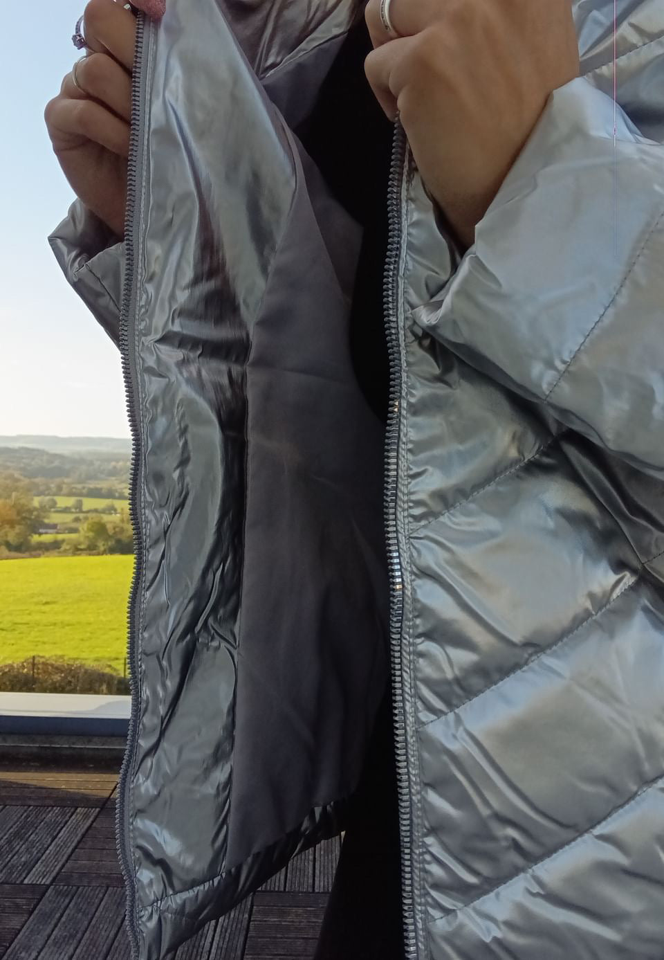 Woman wearing a shiny silver Laura Jo raincoat, unzipped outdoors with scenic countryside view