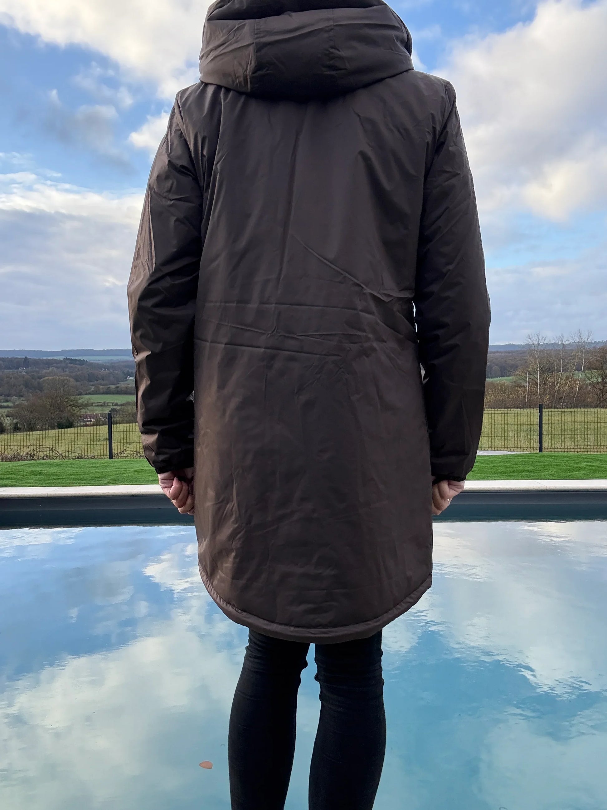 Vue de dos d'une parka longue femme imperméable marron chocolat avec capuche, portée devant une piscine