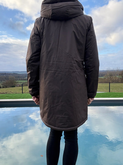 Vue de dos d'une parka longue femme imperméable marron chocolat avec capuche, portée devant une piscine