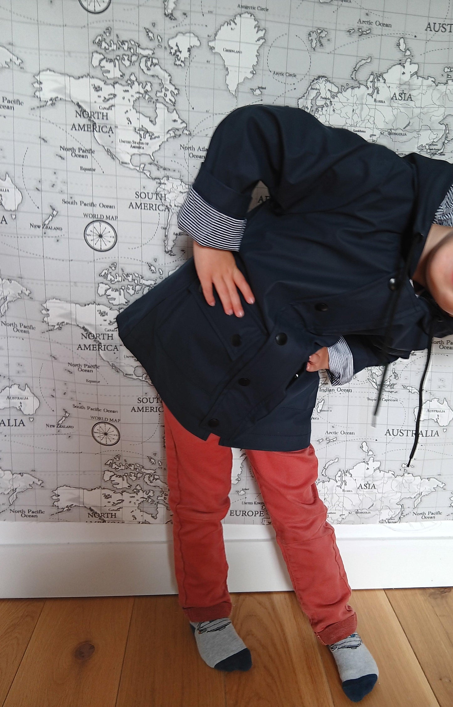 Child in navy raincoat and red pants posing in front of world map wall, Le Petit Placard