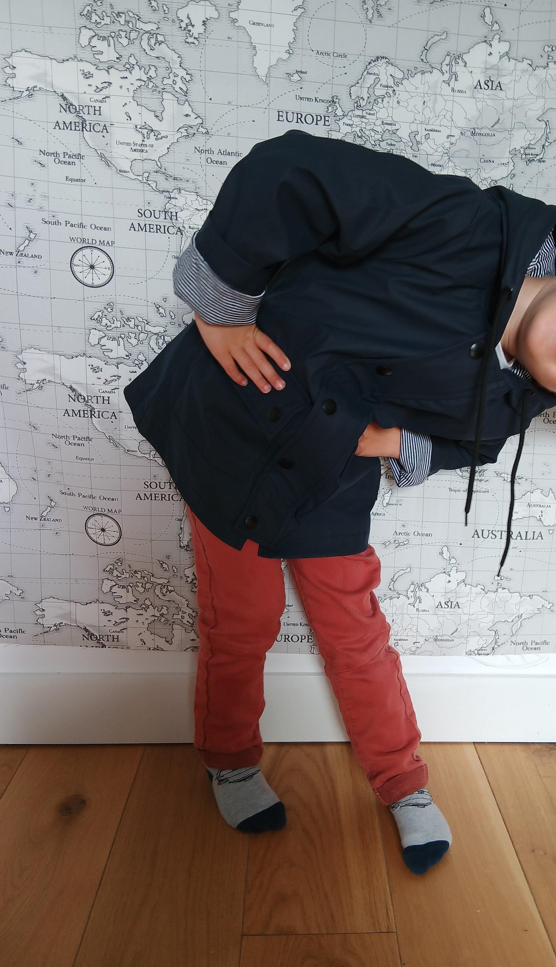 Child in navy blue raincoat and red pants posing in front of world map wall, Le Petit Placard