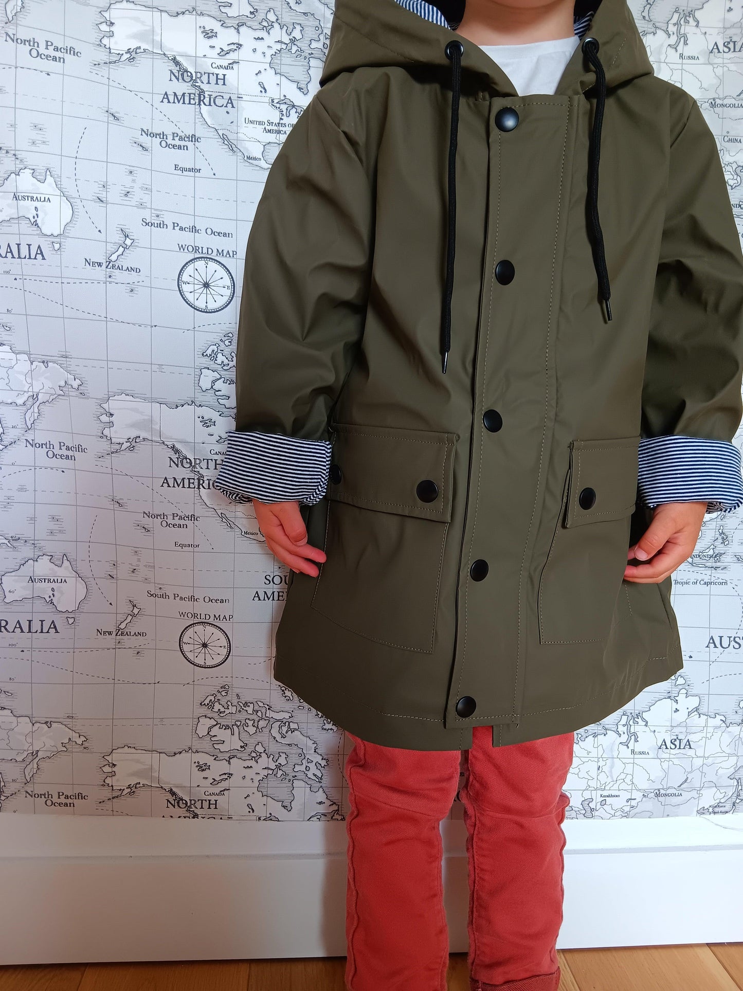 Child wearing khaki raincoat with striped lining, Le Petit Placard, in front of world map wall