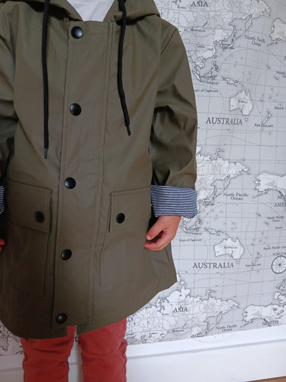 Child wearing khaki raincoat with striped cuffs in front of world map wall, Le Petit Placard
