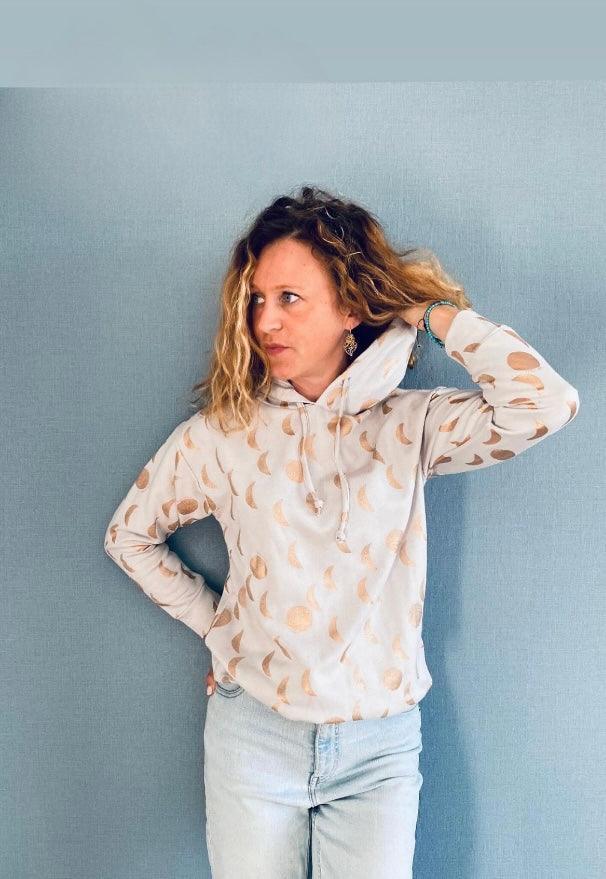 Femme portant un sweat à capuche blanc crème à motifs lune doré rosé et cordon blanc, associé à un jean clair