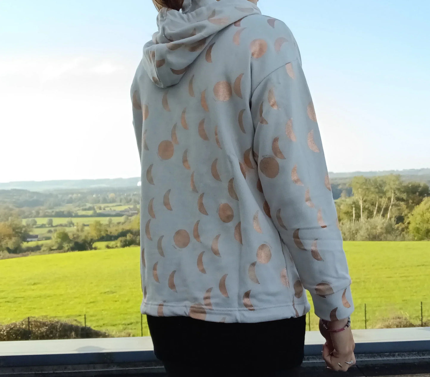 Sweat à capuche blanc crème motifs lune doré rosé porté de dos, vue sur paysage verdoyant