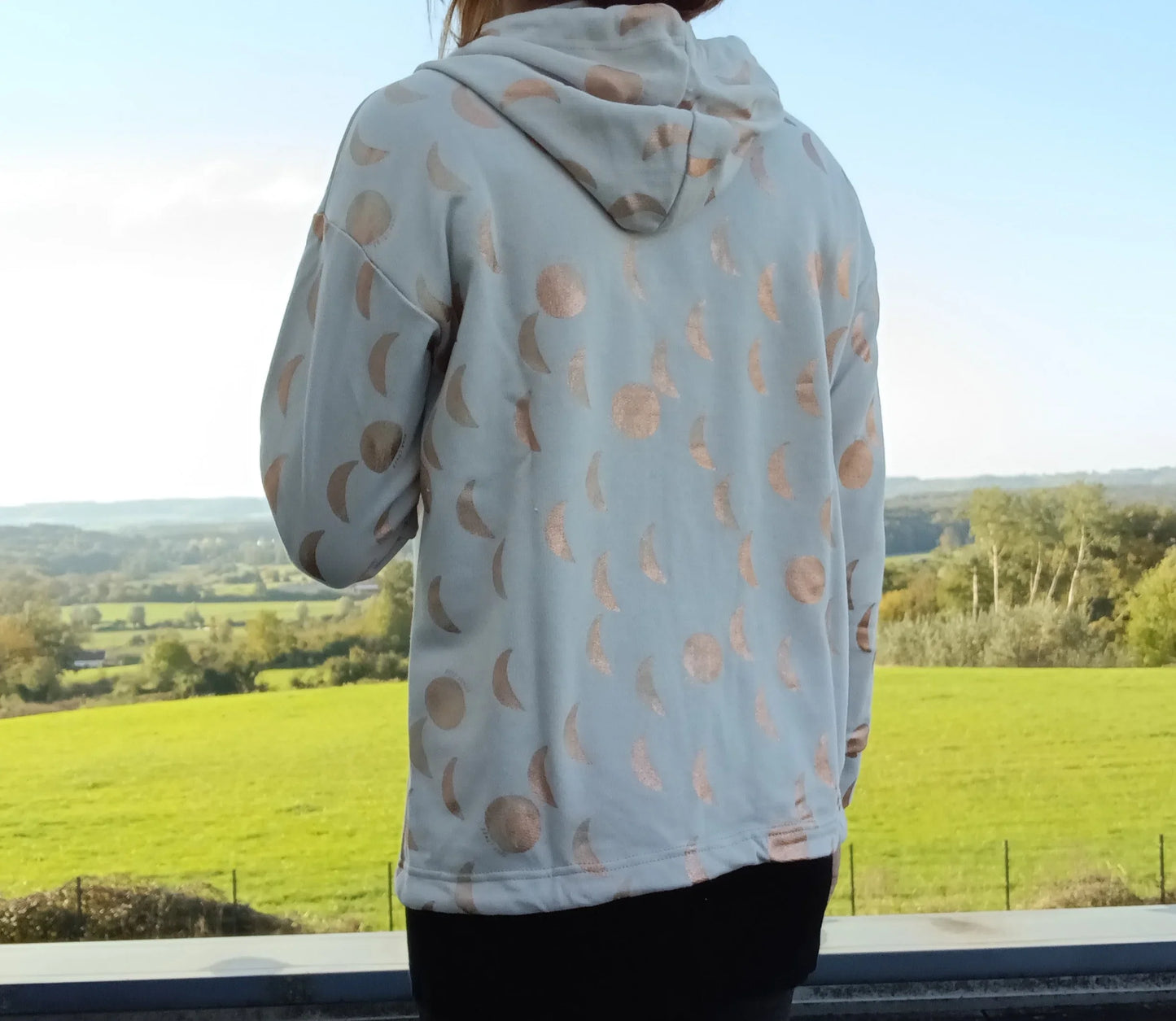 Sweat à capuche blanc crème motifs lune doré rosé, vue de dos, porté par une femme face à un paysage verdoyant