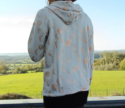 Sweat à capuche blanc crème motifs lune doré rosé, vue de dos, porté par une femme face à un paysage verdoyant
