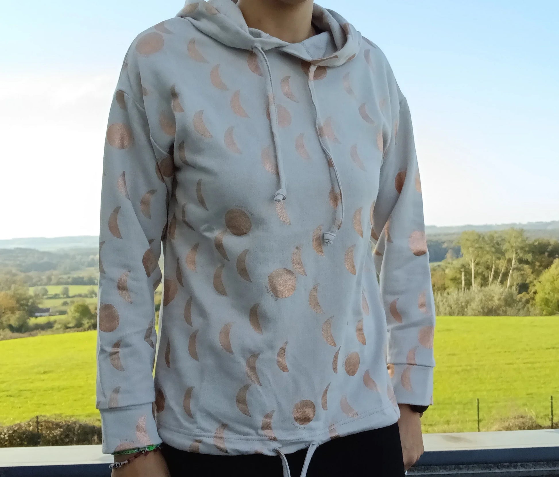Sweat à capuche blanc crème avec cordon, imprimé de motifs lune doré rosé, porté devant un paysage verdoyant