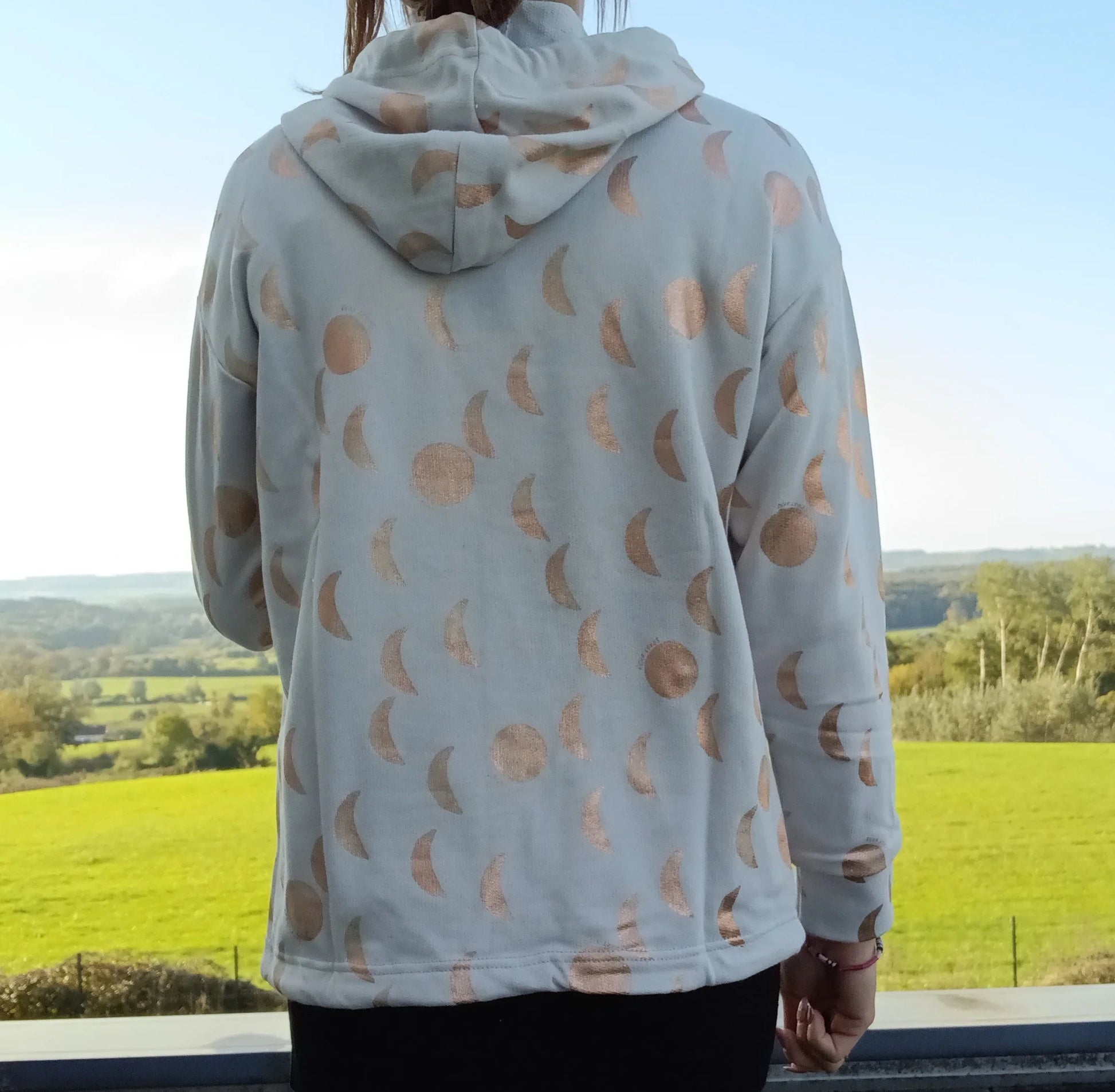 Sweat à capuche blanc crème motifs lune doré rosé, vue de dos, porté par une femme devant un paysage verdoyant