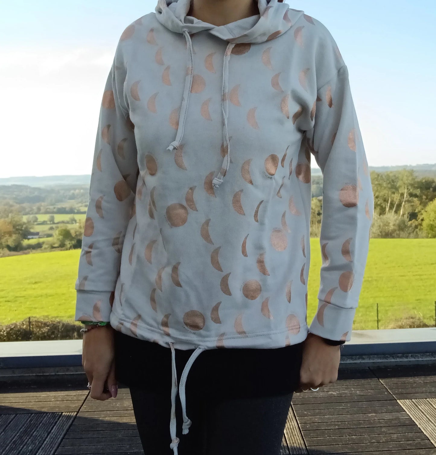 Sweat à capuche blanc crème avec cordon et motifs lune doré rosé, porté par une femme en extérieur