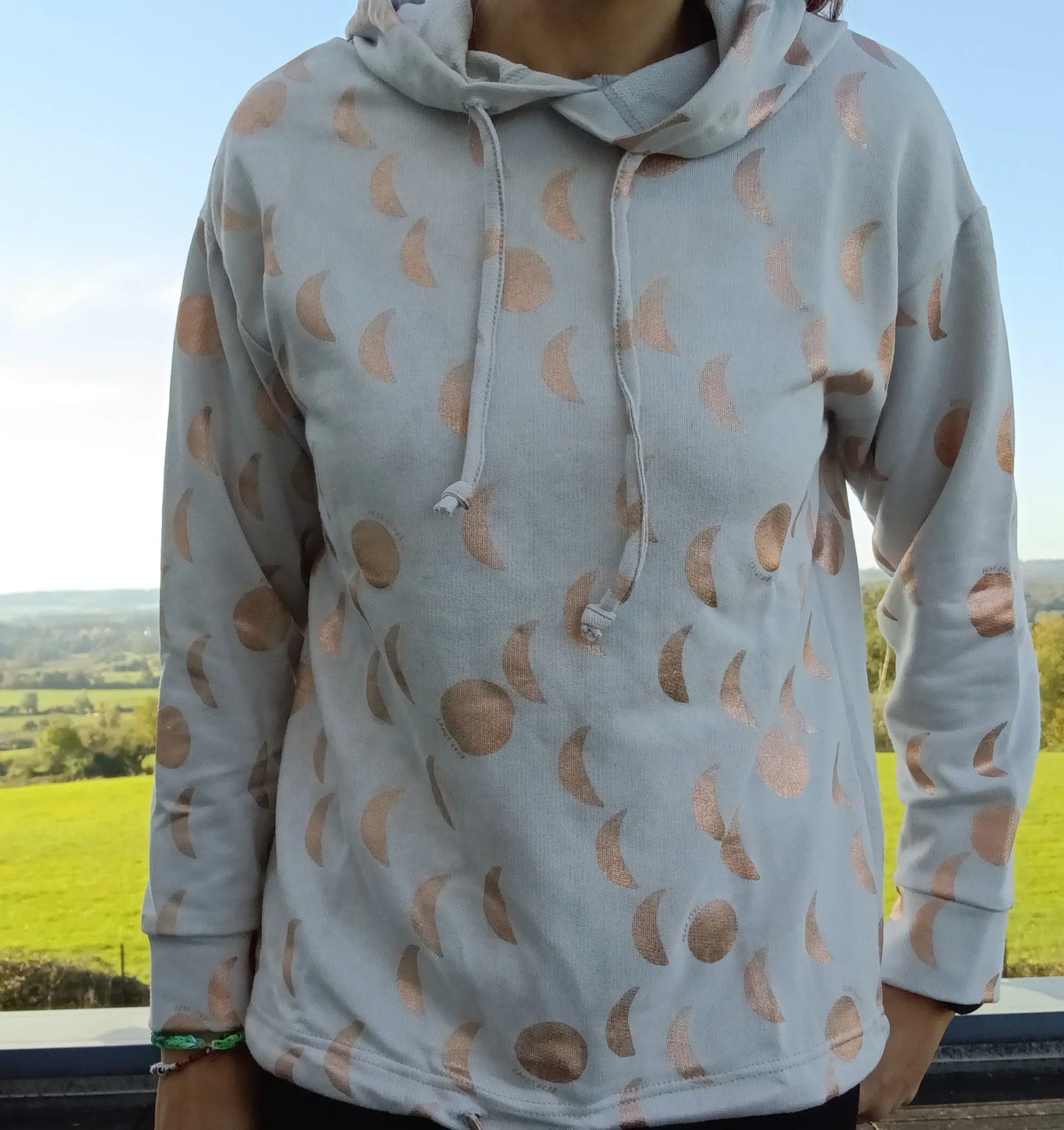 Sweat à capuche blanc crème avec cordon et motifs lune doré rosé, porté par une femme en extérieur