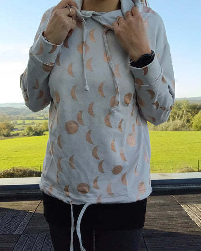 Sweat à capuche blanc crème avec cordon, imprimé motifs lune doré rosé, porté par une femme en extérieur