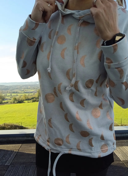 Sweat à capuche blanc crème avec cordon, imprimé motifs lune doré rosé, porté par une femme en extérieur