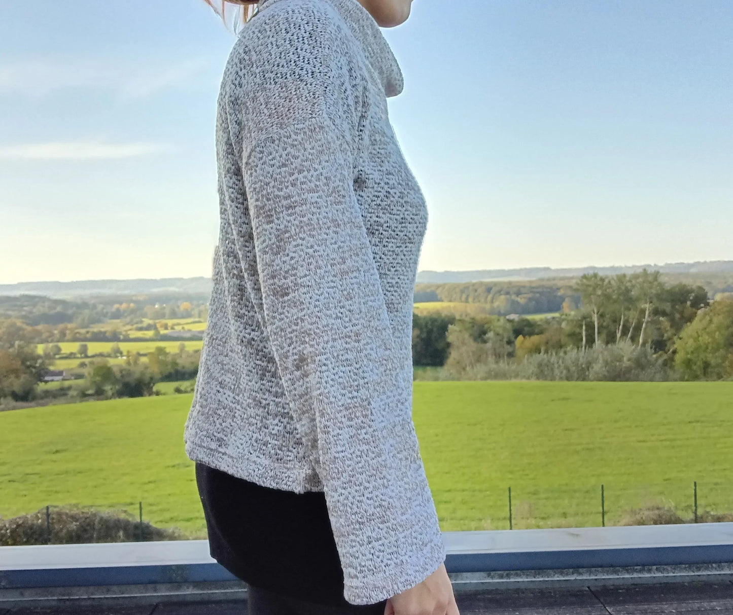 Pull col montant à grosses mailles beige taupe porté de profil par une femme devant un paysage verdoyant