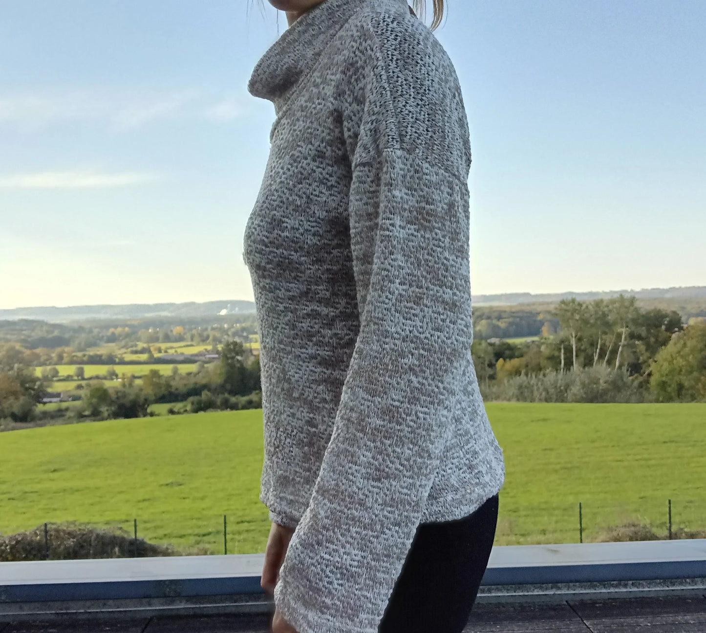 Pull col montant à grosses mailles beige taupe porté de profil par une femme, vue sur campagne verdoyante
