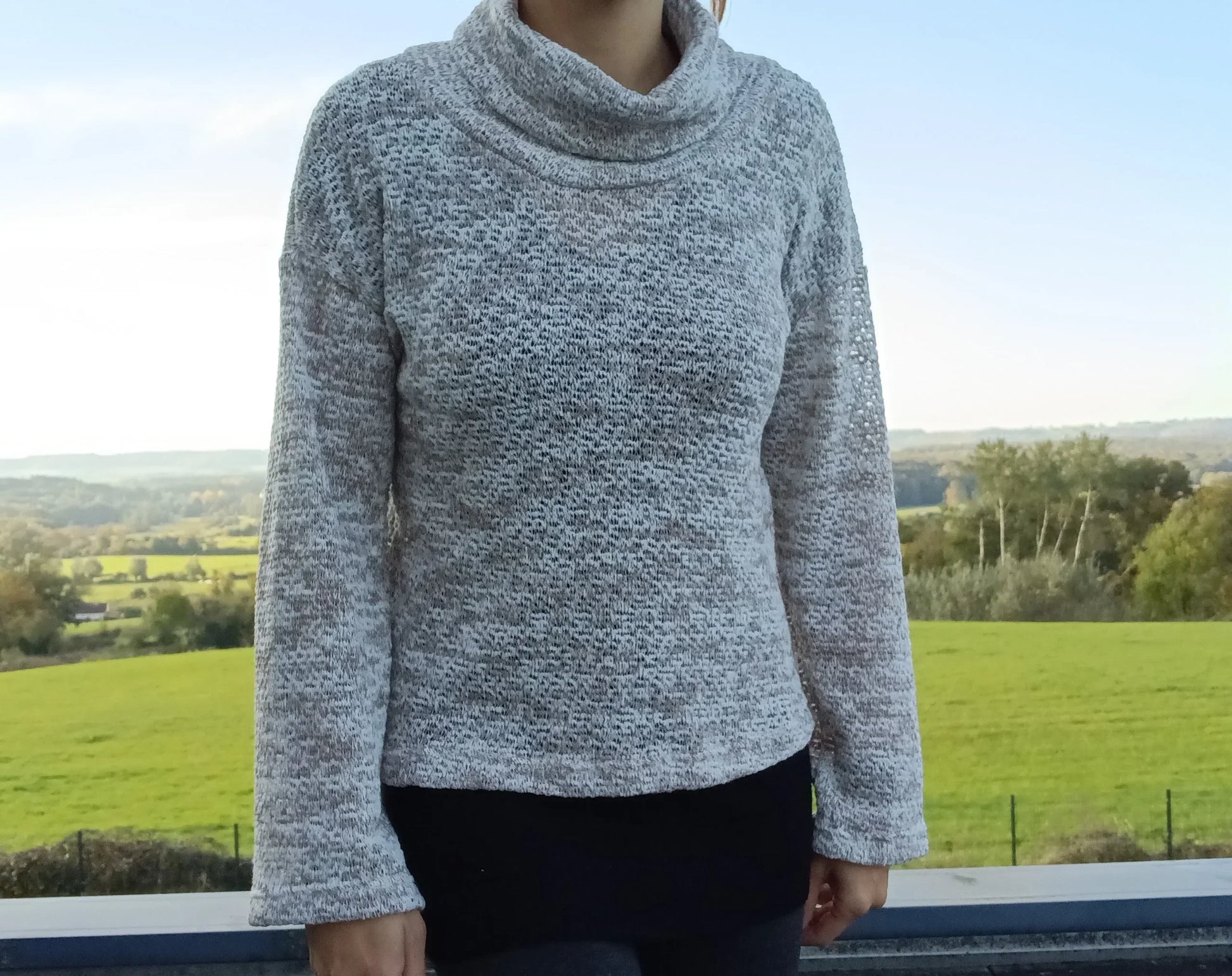 Pull col montant à grosses mailles beige taupe porté par une femme devant un paysage verdoyant