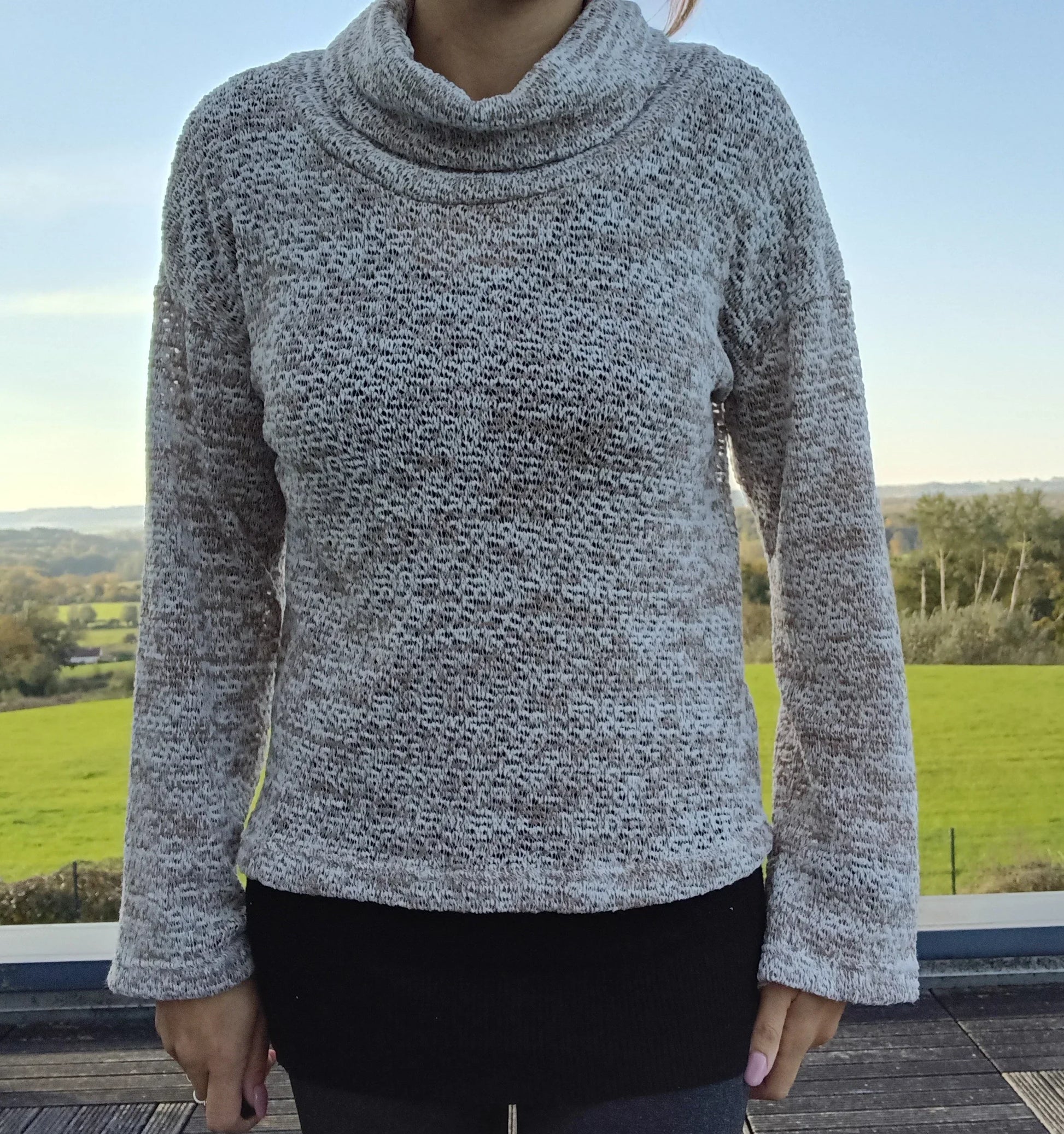 Pull col montant à grosses mailles beige taupe porté par une femme devant un paysage verdoyant