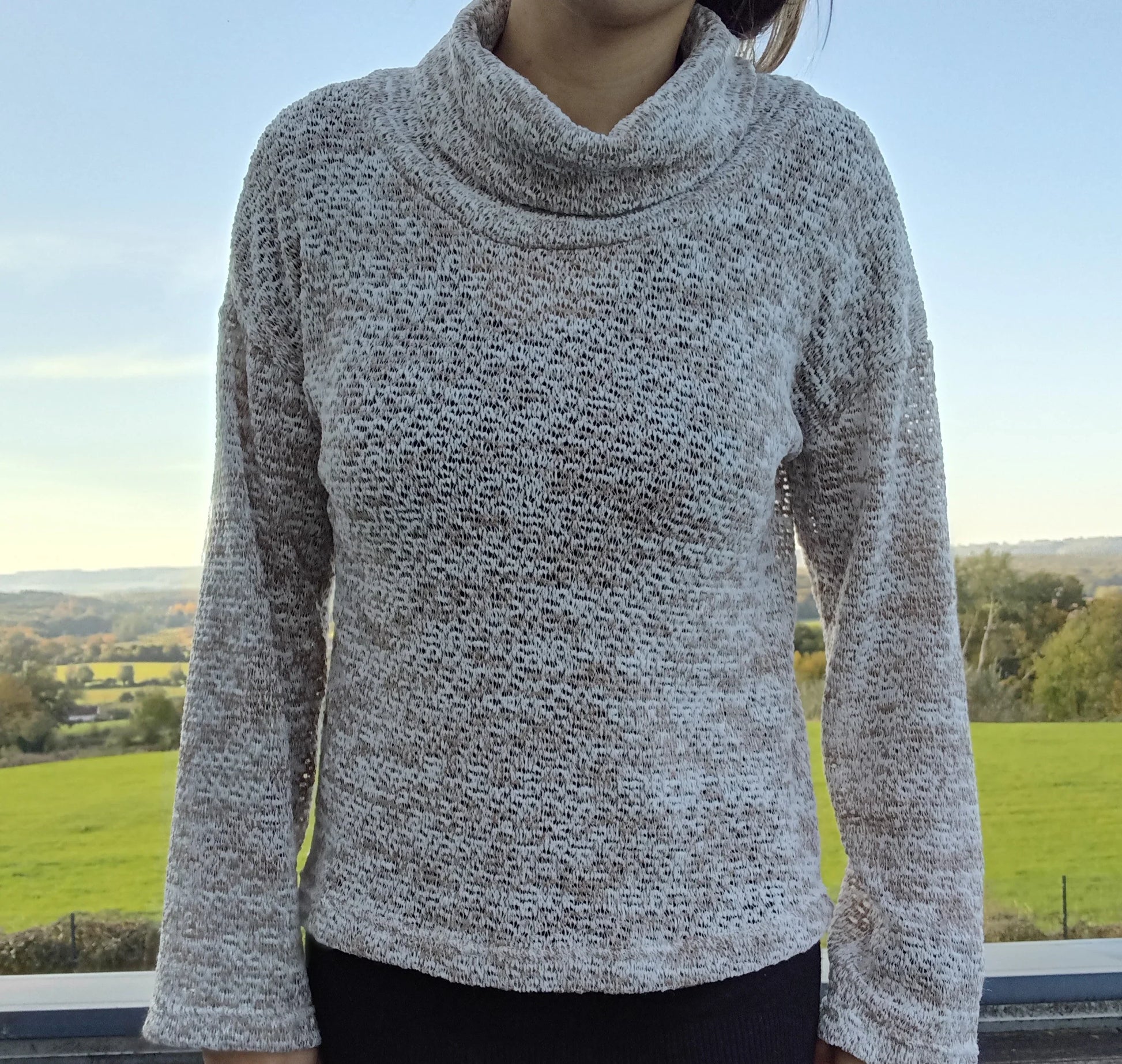 Pull col montant à grosses mailles beige taupe porté par une femme devant un paysage verdoyant