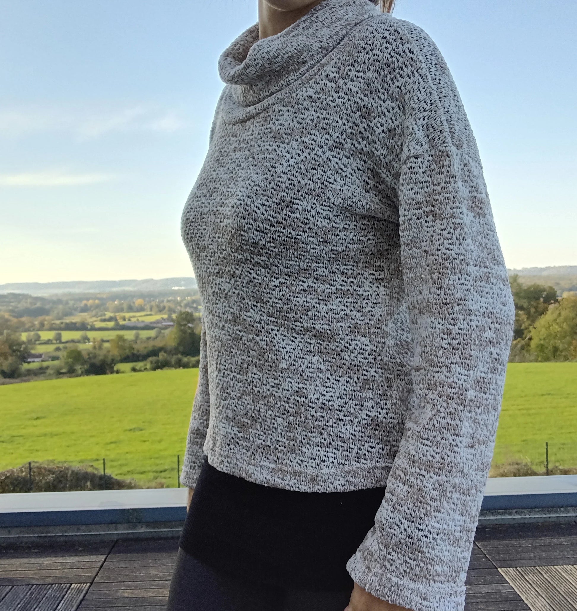 Pull col montant à grosses mailles beige taupe porté de profil, vue extérieure avec paysage verdoyant