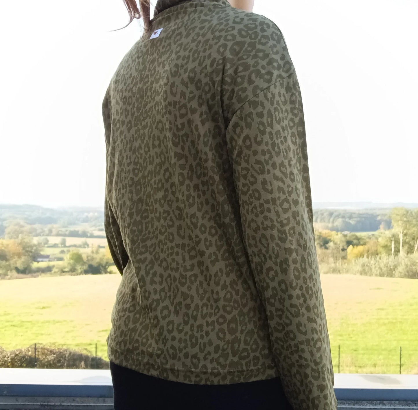 Vue de dos d'un pull fin à col montant motifs léopard kaki porté par une femme face à un paysage verdoyant