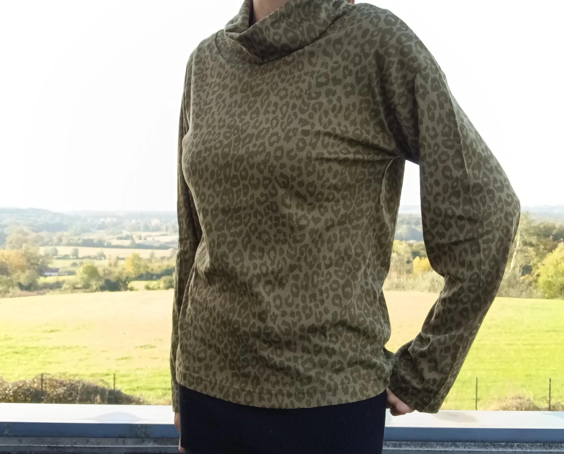 Pull fin à col montant motifs léopard kaki porté par une femme devant un paysage verdoyant
