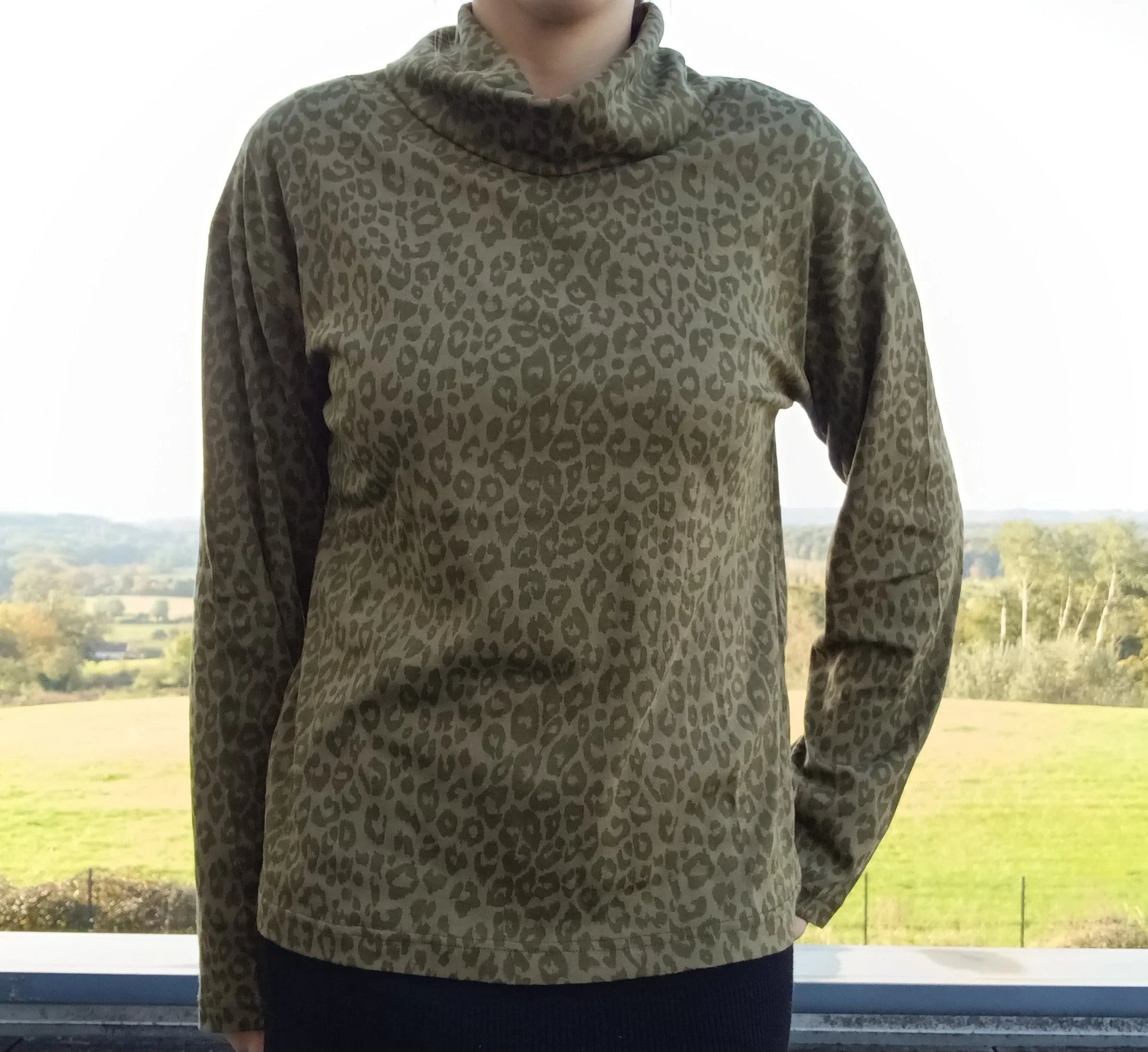 Pull fin à col montant motifs léopard kaki porté par une femme, fond paysage verdoyant