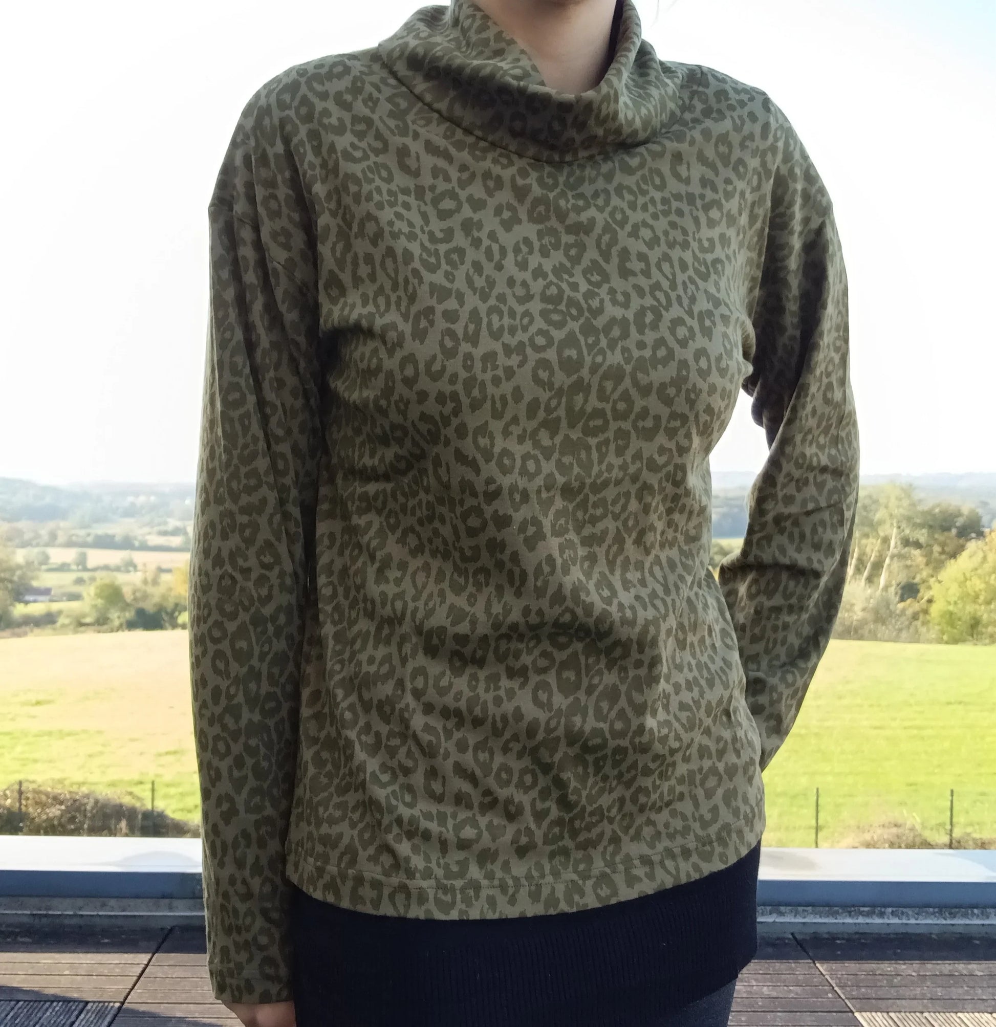 Pull fin à col montant motifs léopard kaki porté par une femme, fond paysage verdoyant