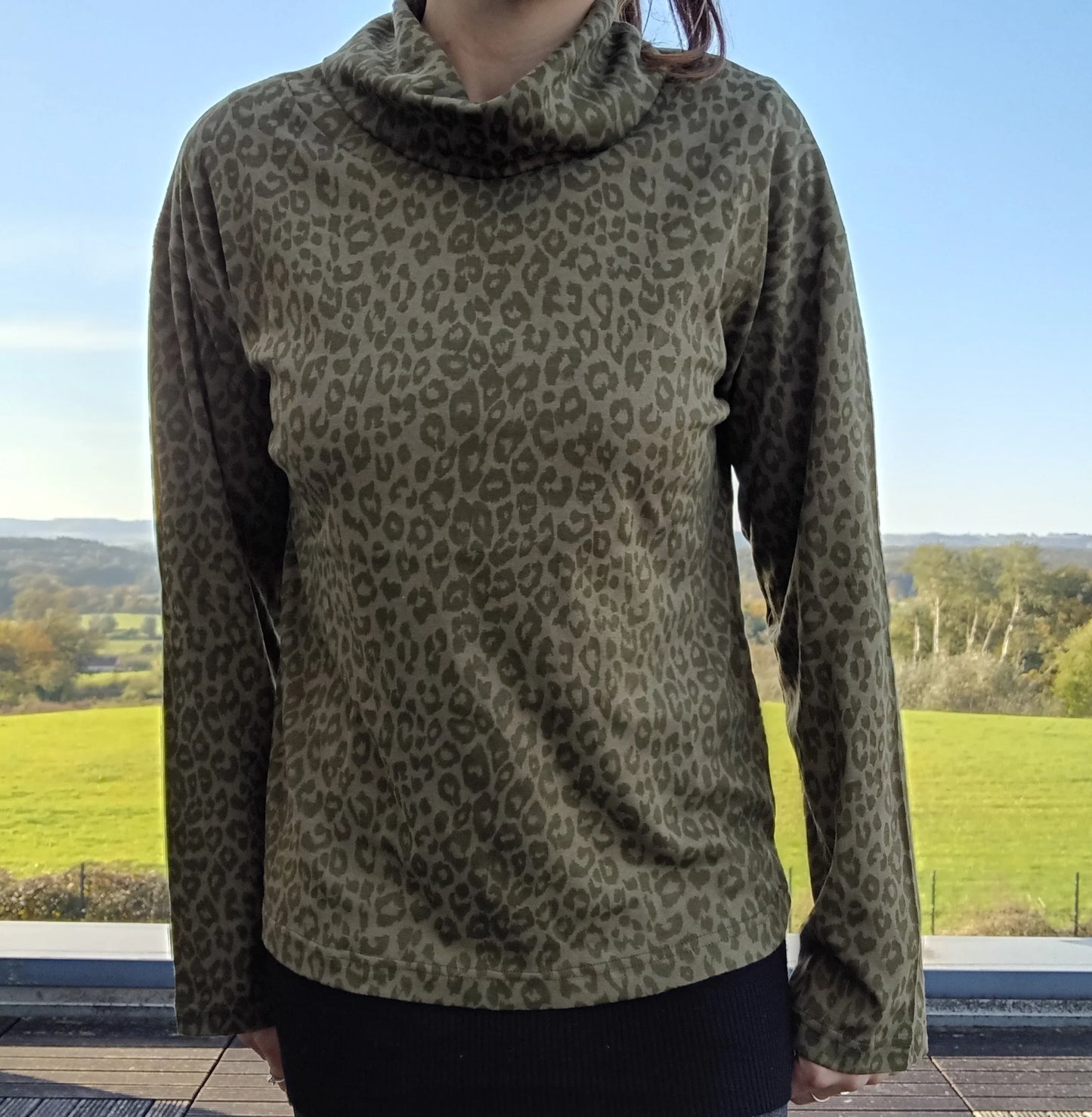 Pull fin à col montant motifs léopard kaki porté par une femme, fond de paysage verdoyant