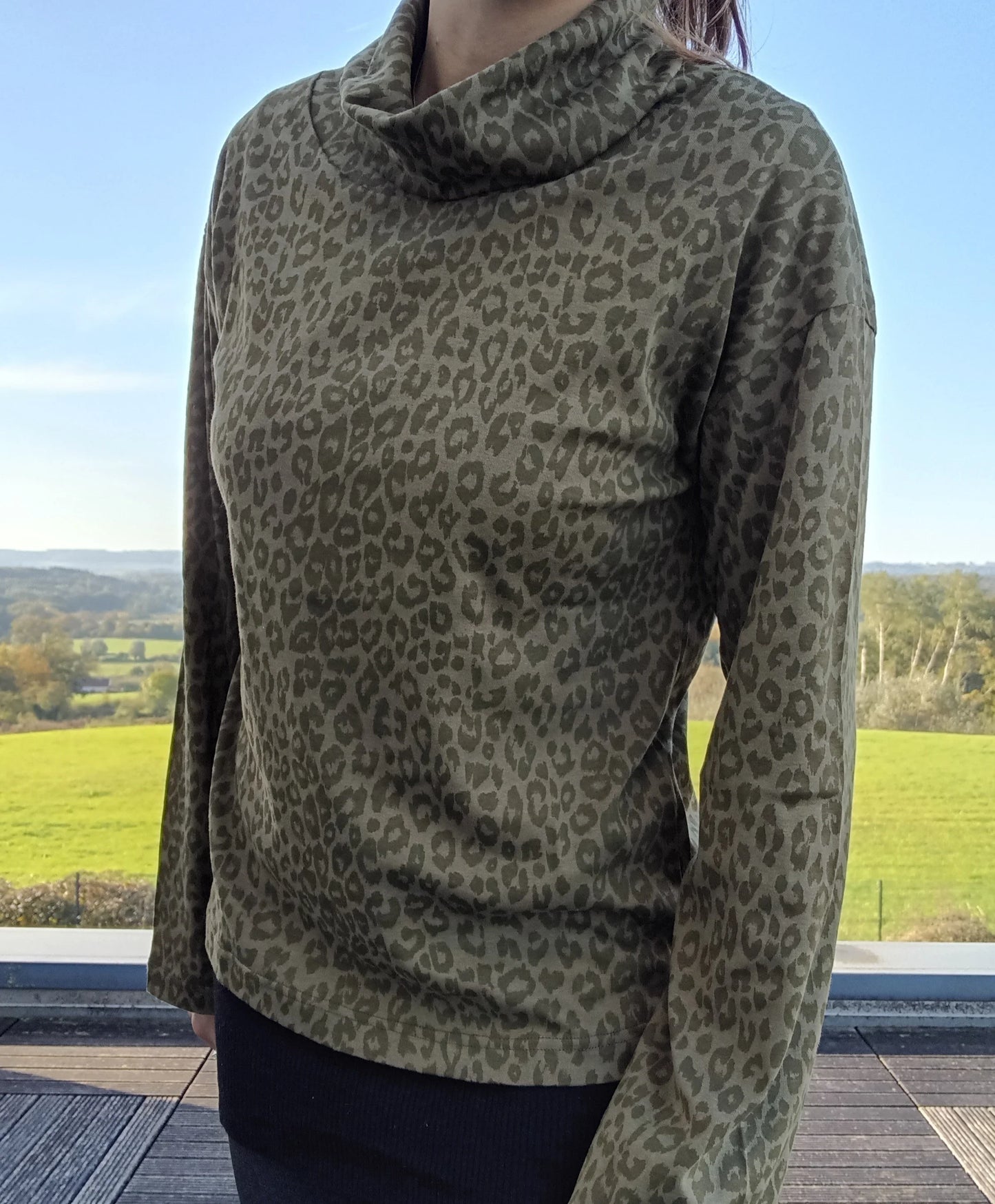 Pull fin à col montant motifs léopard kaki porté par une femme, fond de paysage verdoyant