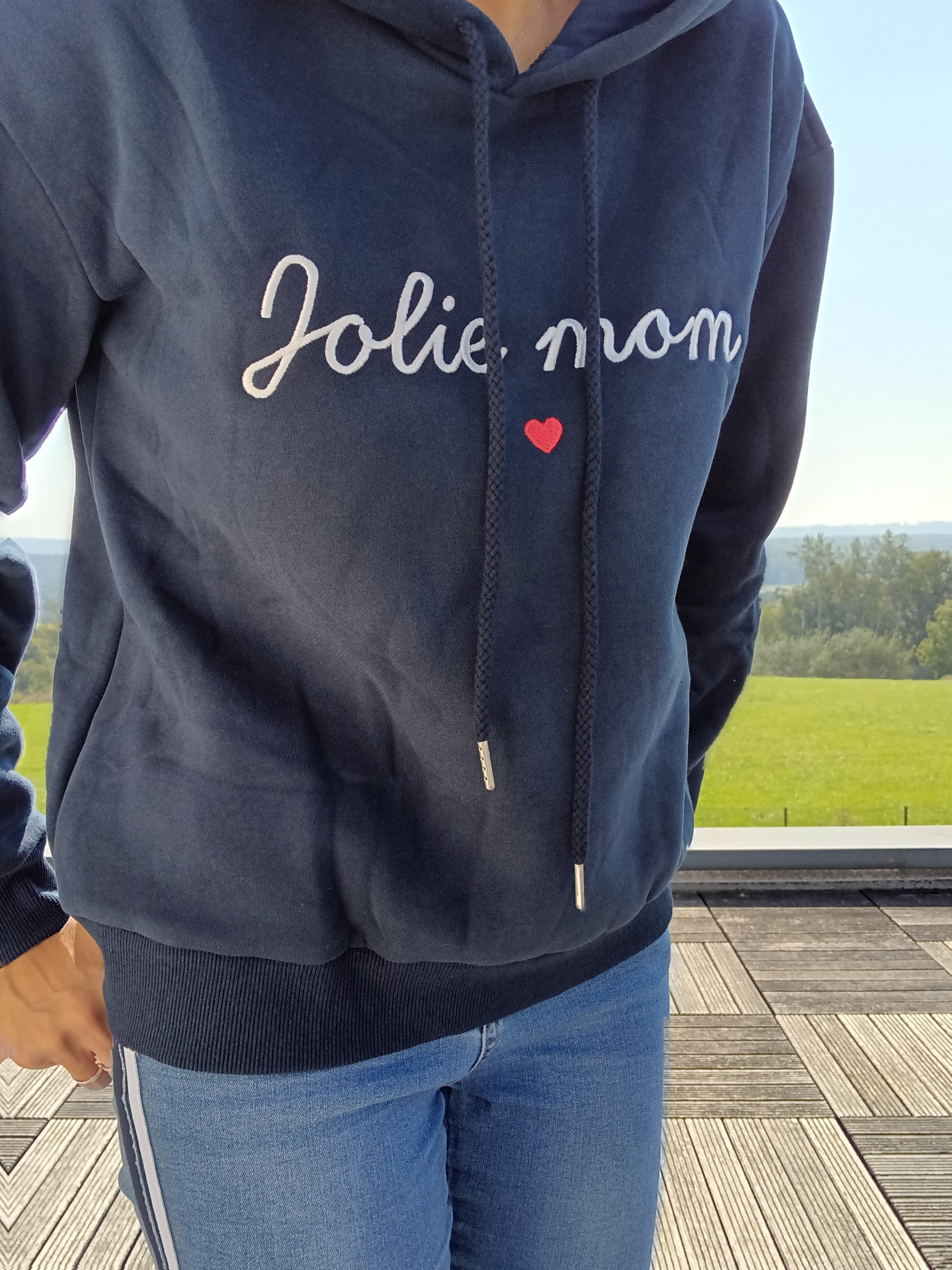 Navy blue hoodie with 'Jolie mom' text and red heart, worn outdoors on a sunny day, Le Petit Placard