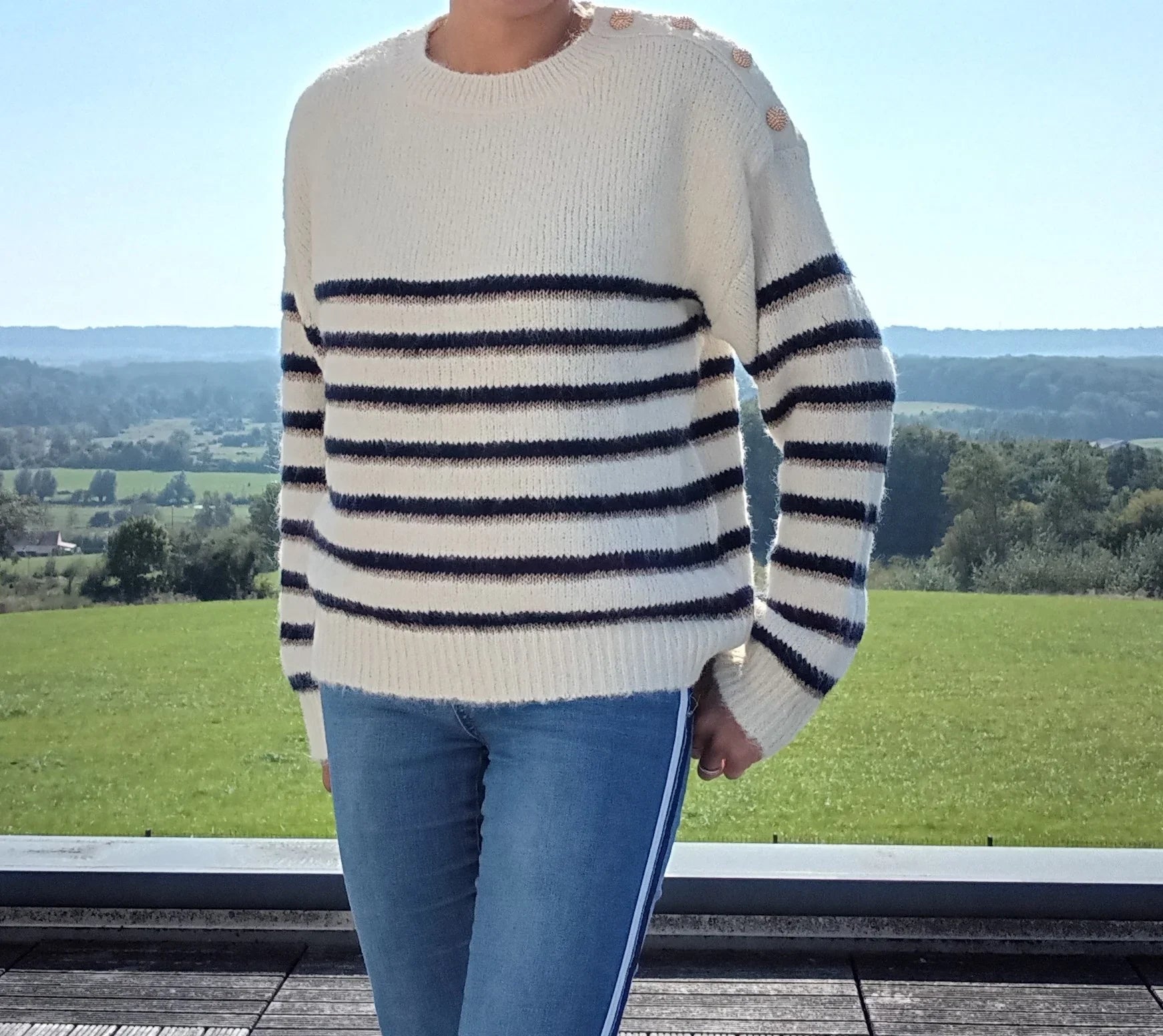 Pull fond blanc rayures bleu marine et dorées avec boutons dorés sur l'épaule, porté avec un jean