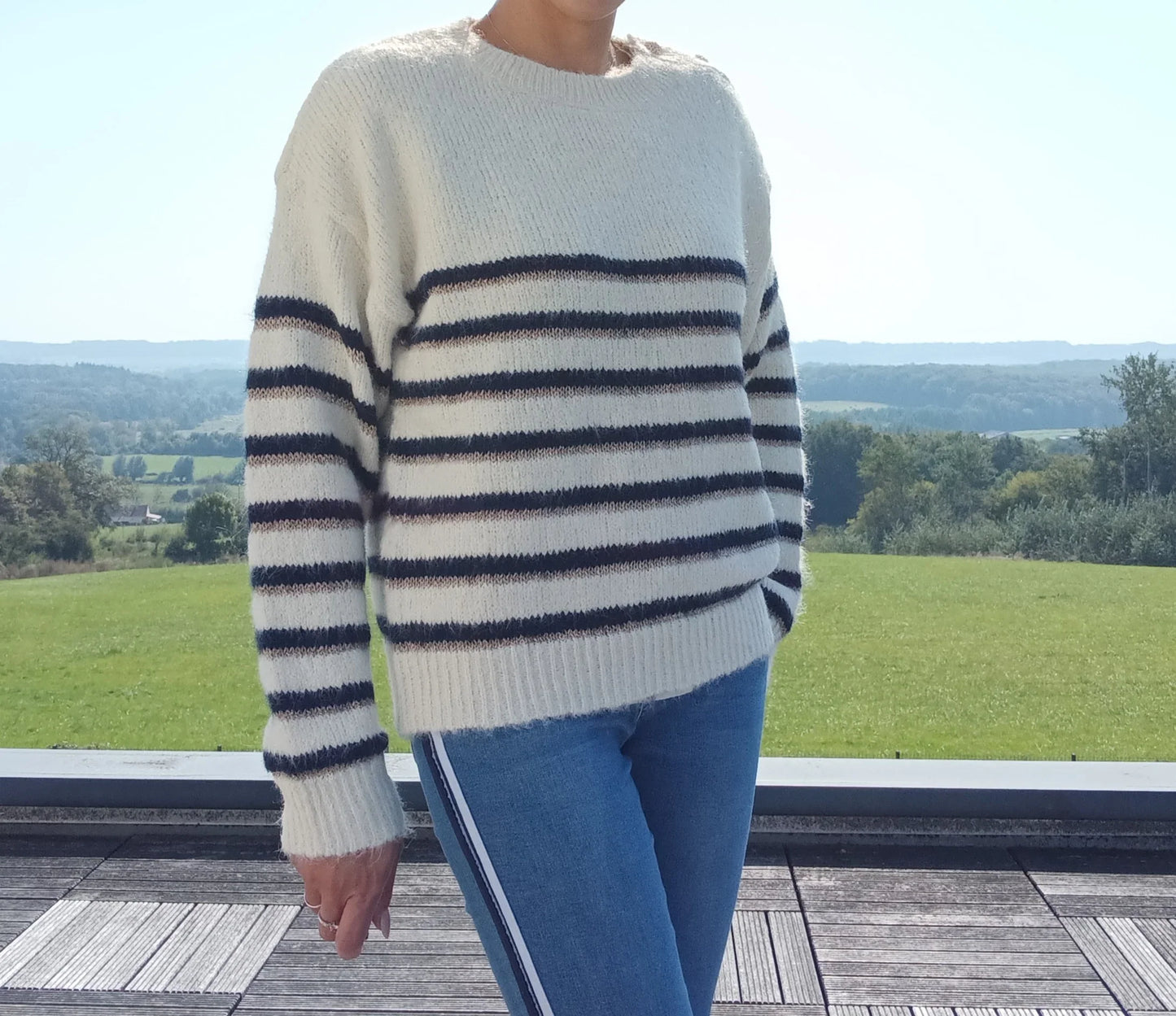 Pull fond blanc double rayures bleu marine et doré porté avec jean, vue extérieure sur terrasse en bois