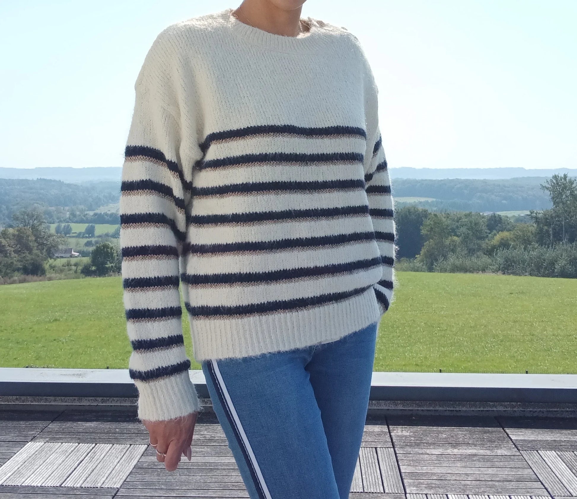 Pull fond blanc double rayures bleu marine et doré porté avec jean, vue extérieure sur terrasse en bois