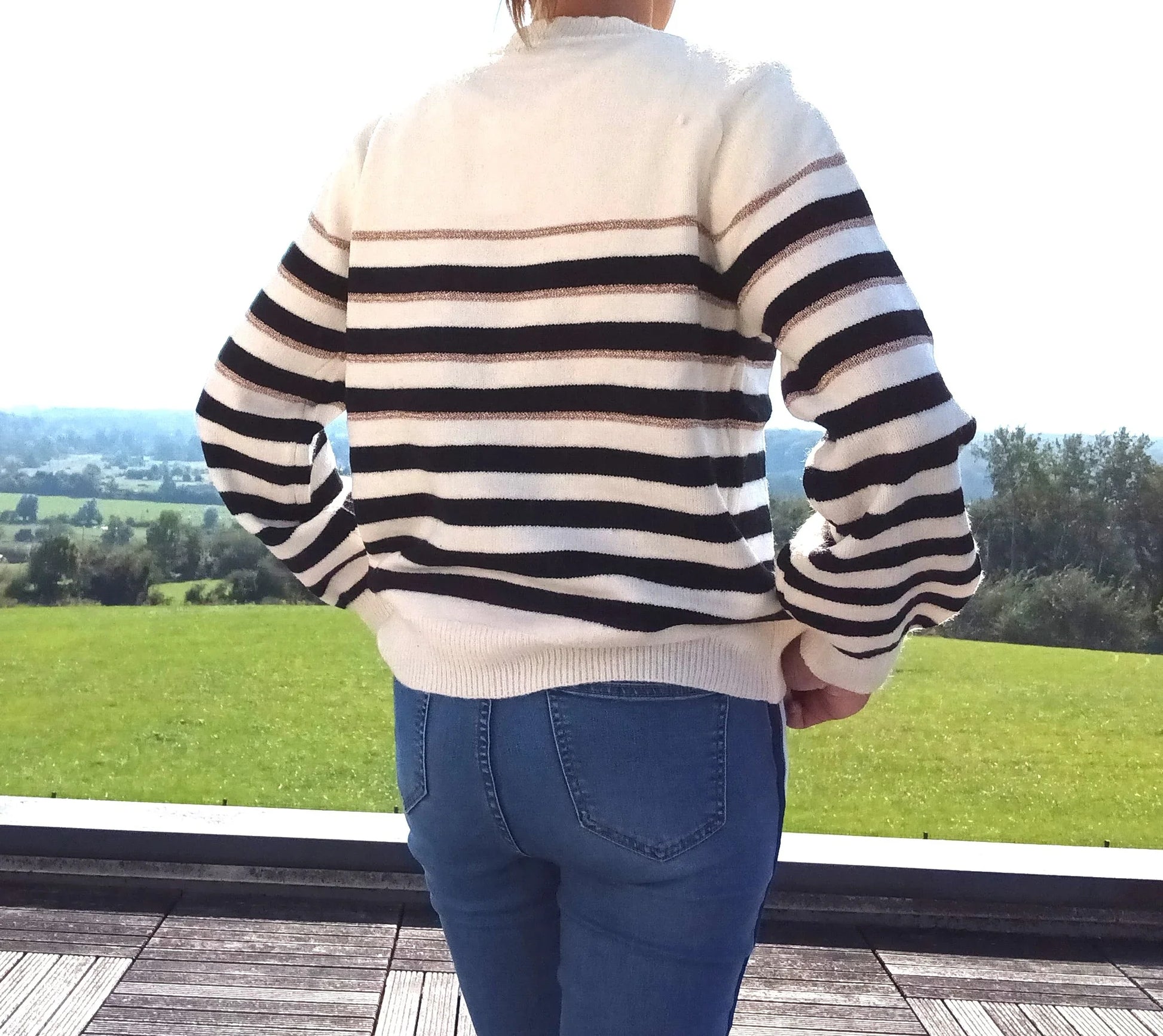 Pull fond blanc larges rayures noires et fines rayures dorées porté de dos avec un jean, vue sur jardin verdoyant