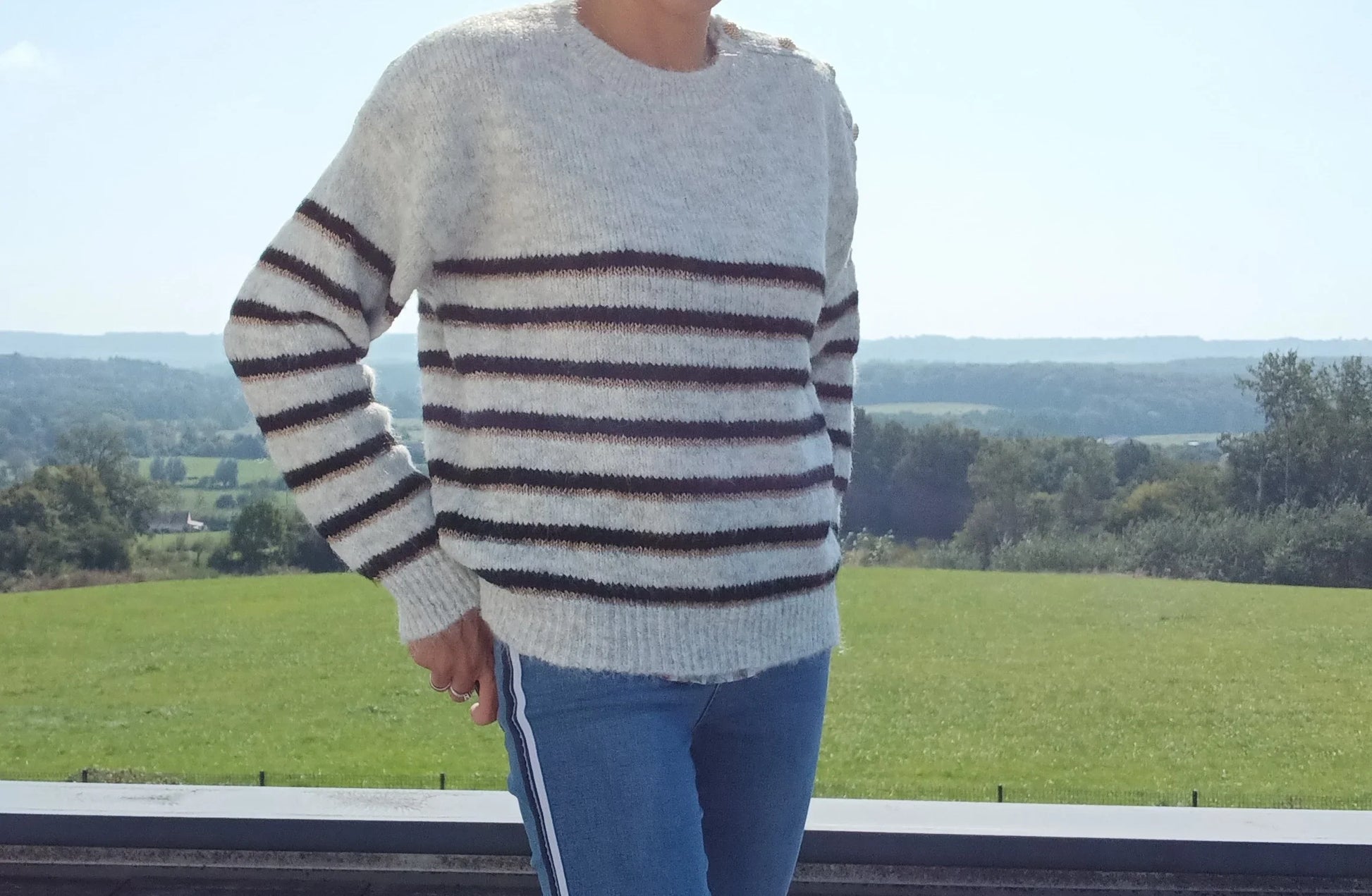 Pull fond gris clair doubles rayures noir et doré porté avec un jean, en extérieur avec vue sur la campagne