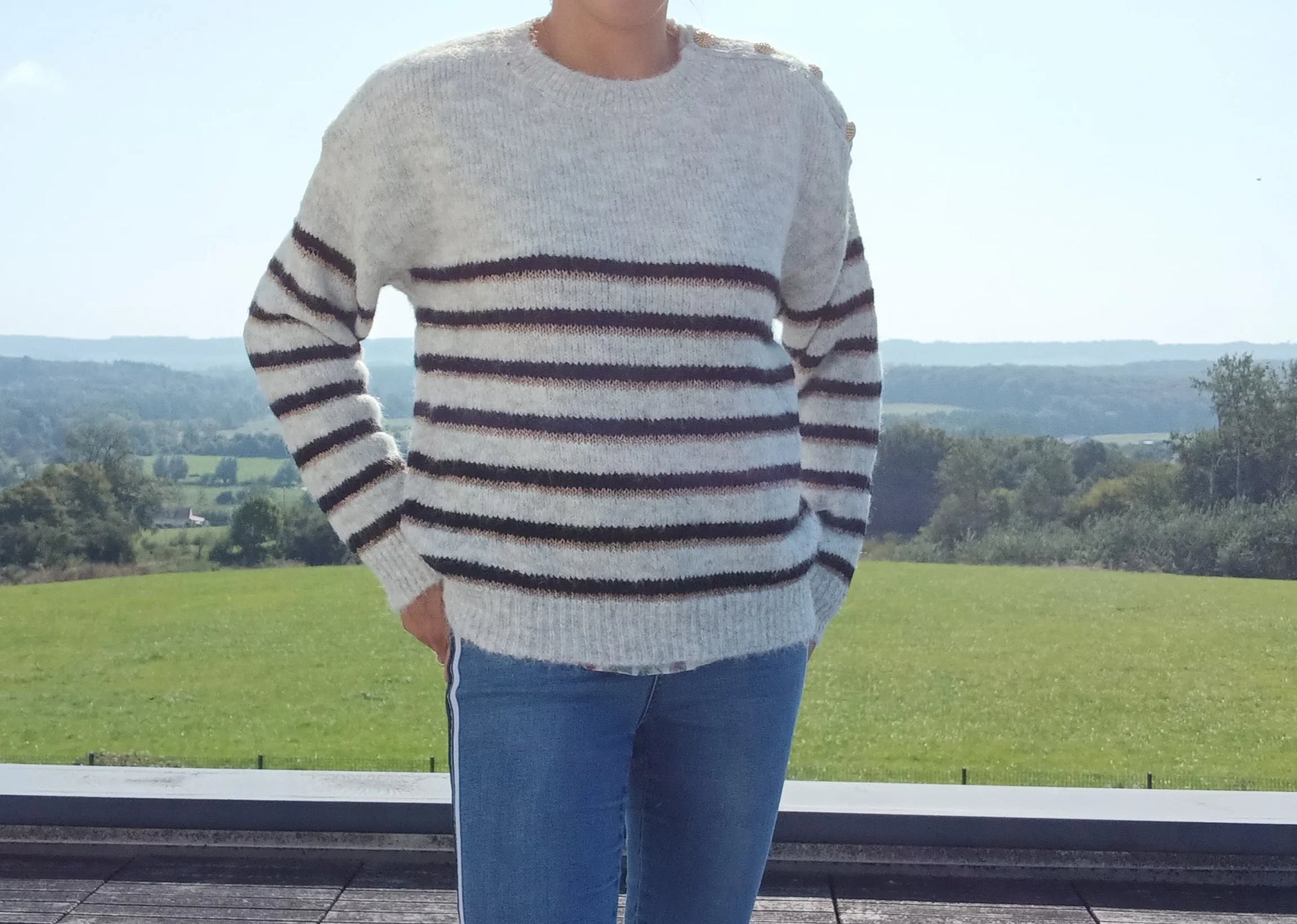 Pull fond gris clair doubles rayures noir et doré porté avec un jean, vue extérieure campagne