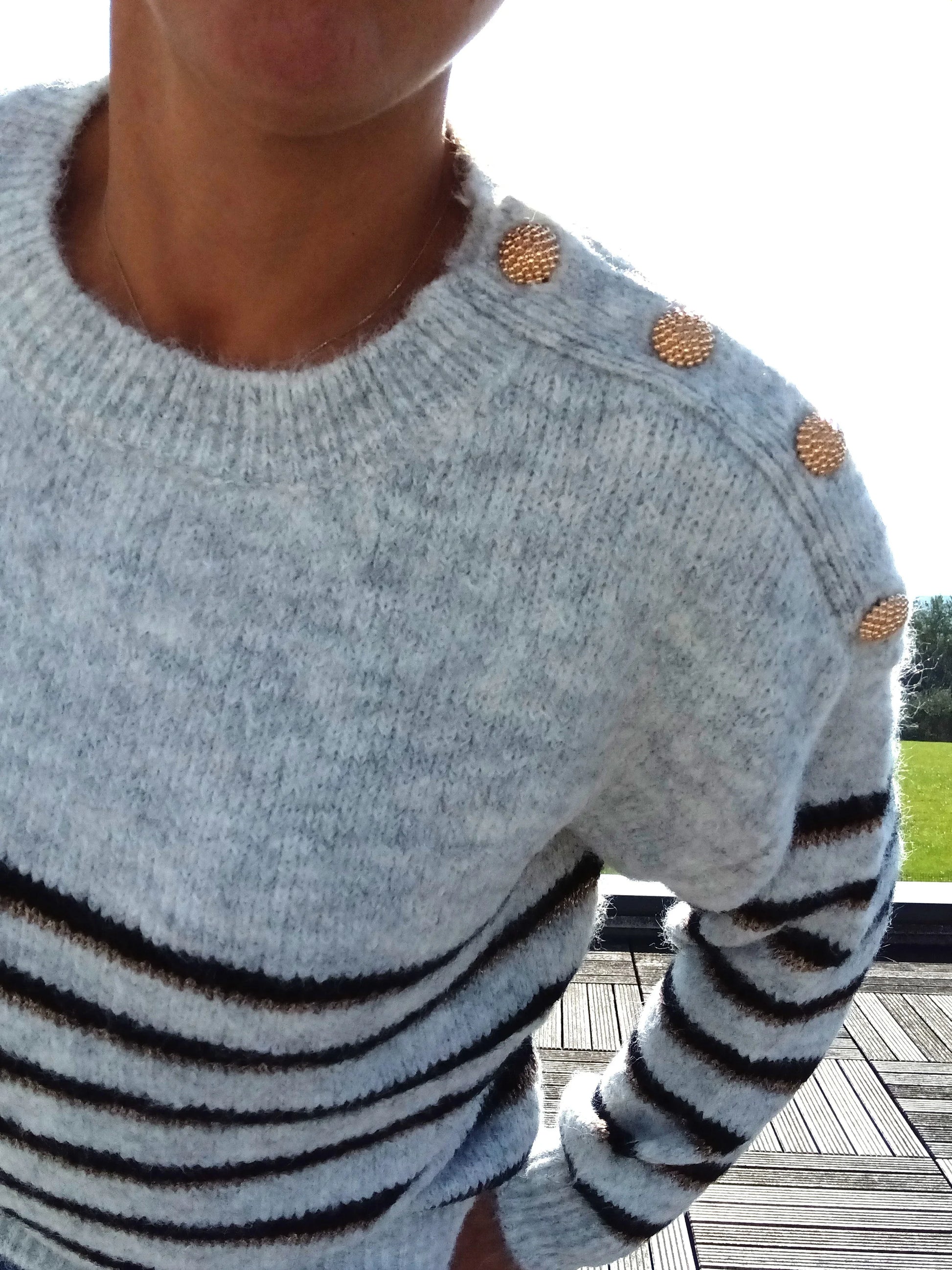 Pull fond gris clair doubles rayures noir et doré porté par une femme, avec boutons dorés sur l'épaule