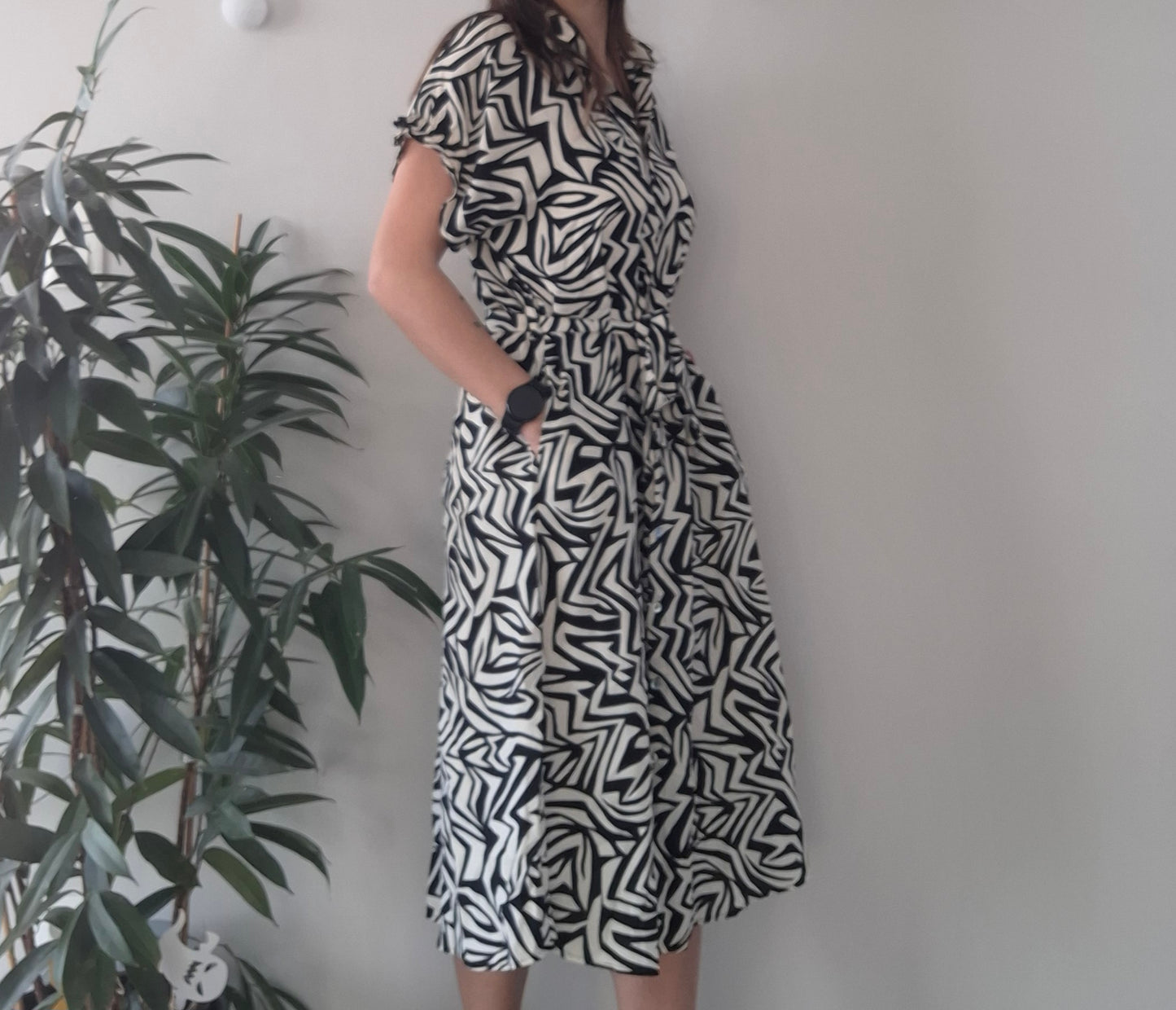 Woman wearing black and white abstract print dress, standing by a plant indoors