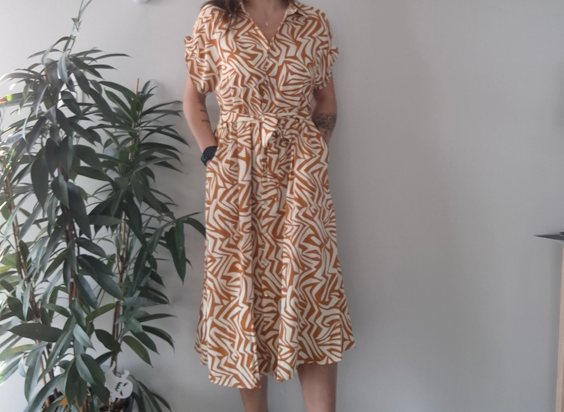 Woman in geometric print dress with short sleeves, standing indoors near a leafy plant