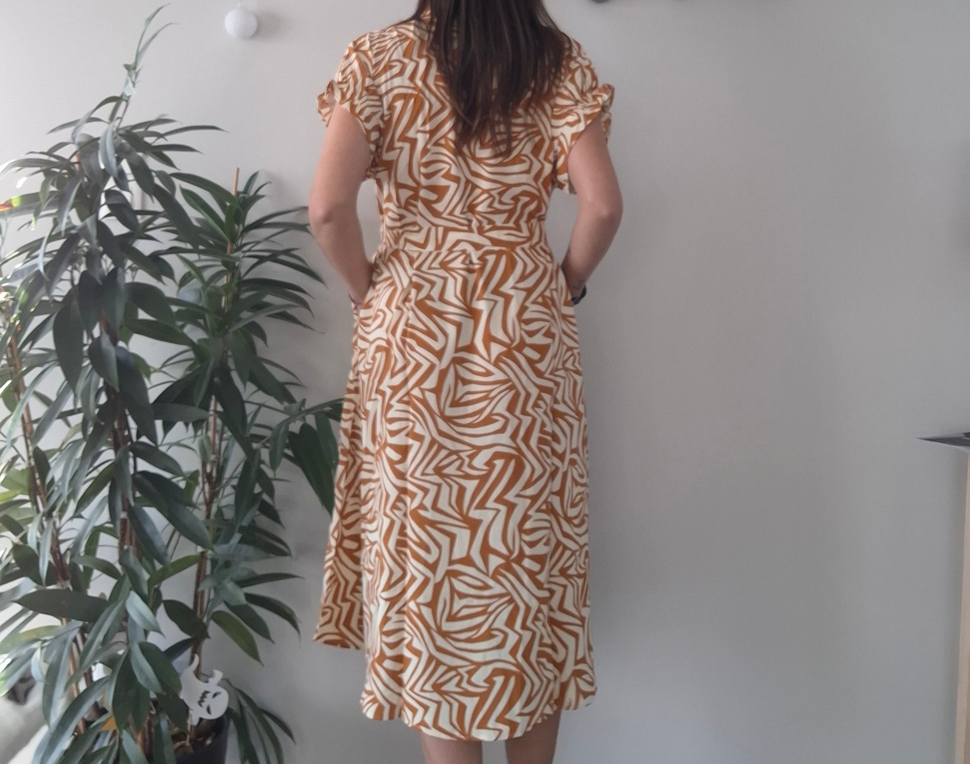 Woman wearing a brown and white geometric print dress, standing indoors near a plant