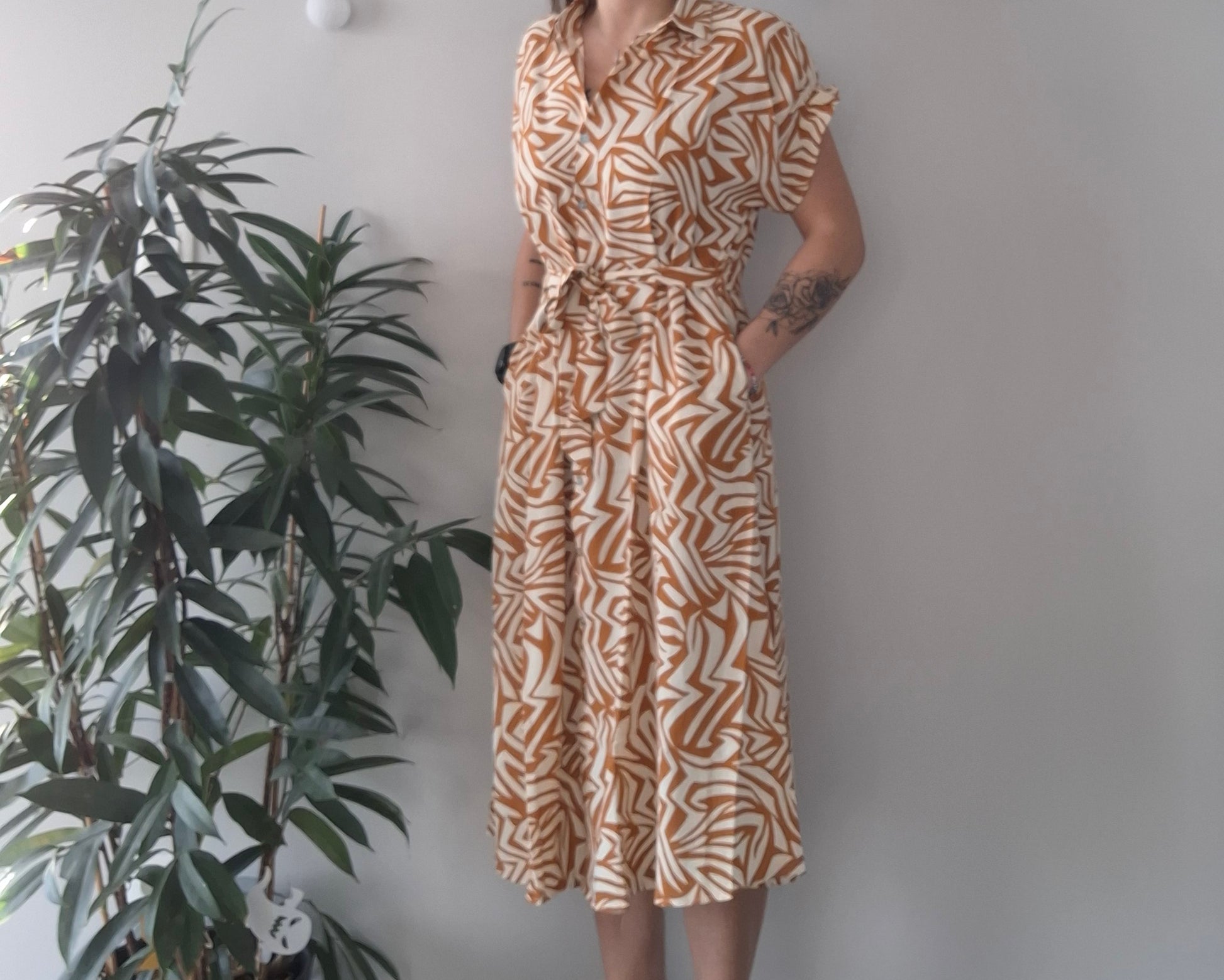 Woman wearing a short-sleeve, brown and white patterned midi dress from Le Petit Placard, standing next to a green indoor plant.