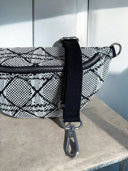 Sac banane effet tissu tressé noir et blanc avec bandoulière noire et fermeture éclair, posé sur une surface en marbre