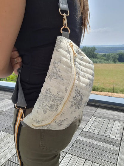 Sac banane en coton matelassé blanc à motif floral gris, avec fermeture éclair dorée et bandoulière gris clair.